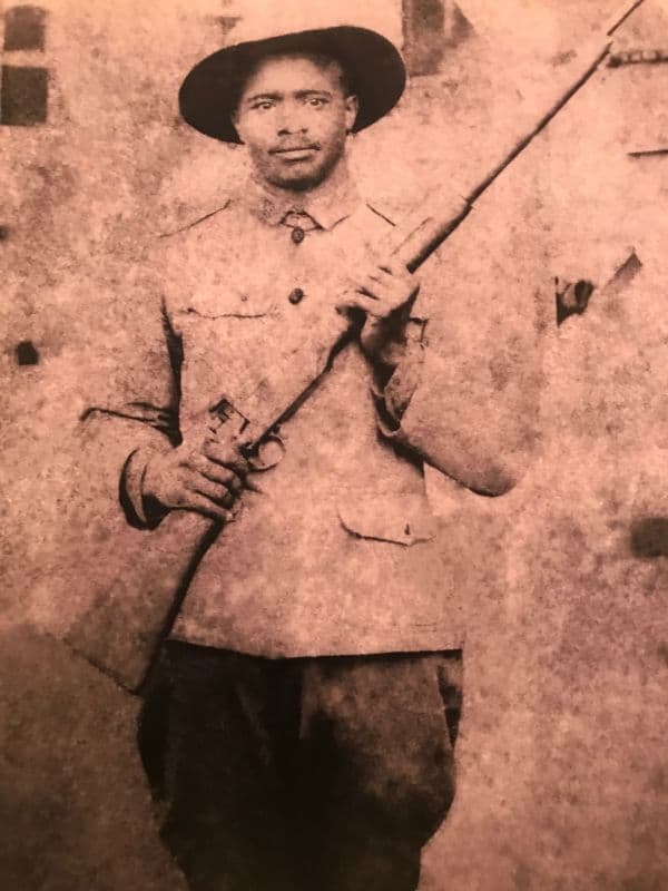 Sepia-toned photograph of a young soldier in a broad-brimmed campaign hat and a service tunic with breast pockets, standing outdoors against a light mottled wall and holding a rifle upright in both hands, muzzle pointing up past his shoulder.