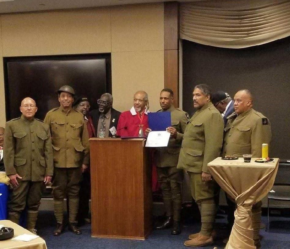 Modern color photograph of a group of men in World War I-pattern service uniforms at an indoor award ceremony, one holding a certificate at a podium.